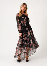 [Color: Black/Rose] A front facing image of a blonde model wearing a special occasion bohemian maxi dress in a black and pink floral print. Designed in chiffon, with a one shoulder detail, a long sleeve, a flowy ruffle trimmed tiered skirt, a high low hemline, an elastic waist, and a smocked elastic neckline. 
