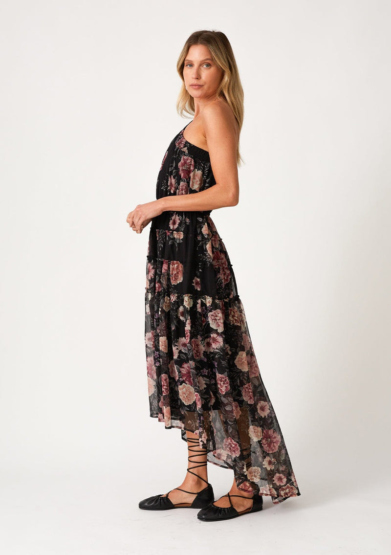 [Color: Black/Rose] A side facing image of a blonde model wearing a special occasion bohemian maxi dress in a black and pink floral print. Designed in chiffon, with a one shoulder detail, a long sleeve, a flowy ruffle trimmed tiered skirt, a high low hemline, an elastic waist, and a smocked elastic neckline. 