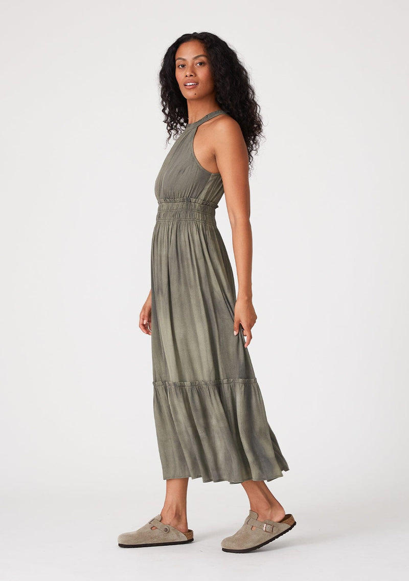 [Color: Olive] A side facing image of a brunette model wearing an olive green bohemian maxi dress in a soft pink vintage wash. With a ruffle trimmed halter neckline, a smocked elastic waist, a tiered long skirt, and a back keyhole detail with an adjustable tie.