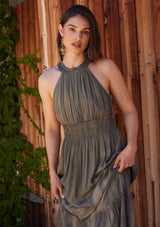 [Color: Olive] A close up front facing image of a brunette model standing outside wearing an olive green bohemian maxi dress in a soft pink vintage wash. With a ruffle trimmed halter neckline, a smocked elastic waist, a tiered long skirt, and a back keyhole detail with an adjustable tie.