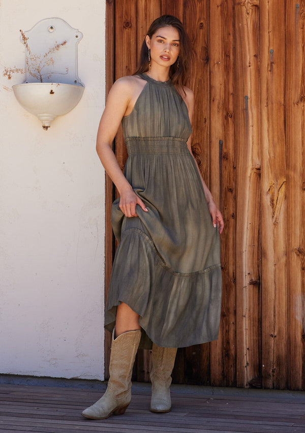 [Color: Olive] A front facing image of a brunette model standing outside wearing an olive green bohemian maxi dress in a soft pink vintage wash. With a ruffle trimmed halter neckline, a smocked elastic waist, a tiered long skirt, and a back keyhole detail with an adjustable tie.
