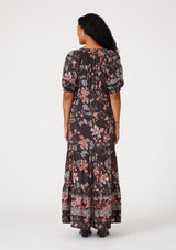 [Color: Brown/Dusty Lilac] A back facing image of a brunette model wearing a bohemian fall maxi dress in a brown and dusty purple floral print. With short puff sleeves, a v neckline, a long tiered skirt, side pockets, and a drawstring waist with tassel ties. 