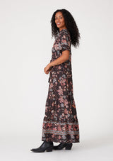 [Color: Brown/Dusty Lilac] A side facing image of a brunette model wearing a bohemian fall maxi dress in a brown and dusty purple floral print. With short puff sleeves, a v neckline, a long tiered skirt, side pockets, and a drawstring waist with tassel ties. 
