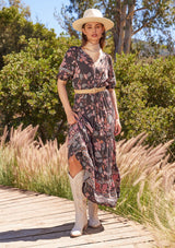 [Color: Brown/Dusty Lilac] A front facing image of a brunette model standing outside wearing a bohemian fall maxi dress in a brown and dusty purple floral print. With short puff sleeves, a v neckline, a long tiered skirt, side pockets, and a drawstring waist with tassel ties.