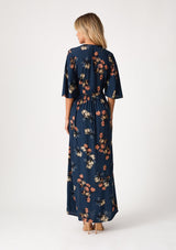 [Color: Teal/Dusty Blush] A back facing image of a blonde model wearing a bohemian fall maxi dress in a teal blue floral print. With half length sleeves, a deep v neckline, an empire waist, a long flowy tiered skirt, and a side slit. 