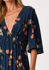 [Color: Teal/Dusty Blush] A close up front facing image of a blonde model wearing a bohemian fall maxi dress in a teal blue floral print. With half length sleeves, a deep v neckline, an empire waist, a long flowy tiered skirt, and a side slit. 