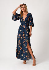 [Color: Teal/Dusty Blush] A front facing image of a blonde model wearing a bohemian fall maxi dress in a teal blue floral print. With half length sleeves, a deep v neckline, an empire waist, a long flowy tiered skirt, and a side slit. 