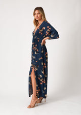 [Color: Teal/Dusty Blush] A side facing image of a blonde model wearing a bohemian fall maxi dress in a teal blue floral print. With half length sleeves, a deep v neckline, an empire waist, a long flowy tiered skirt, and a side slit. 