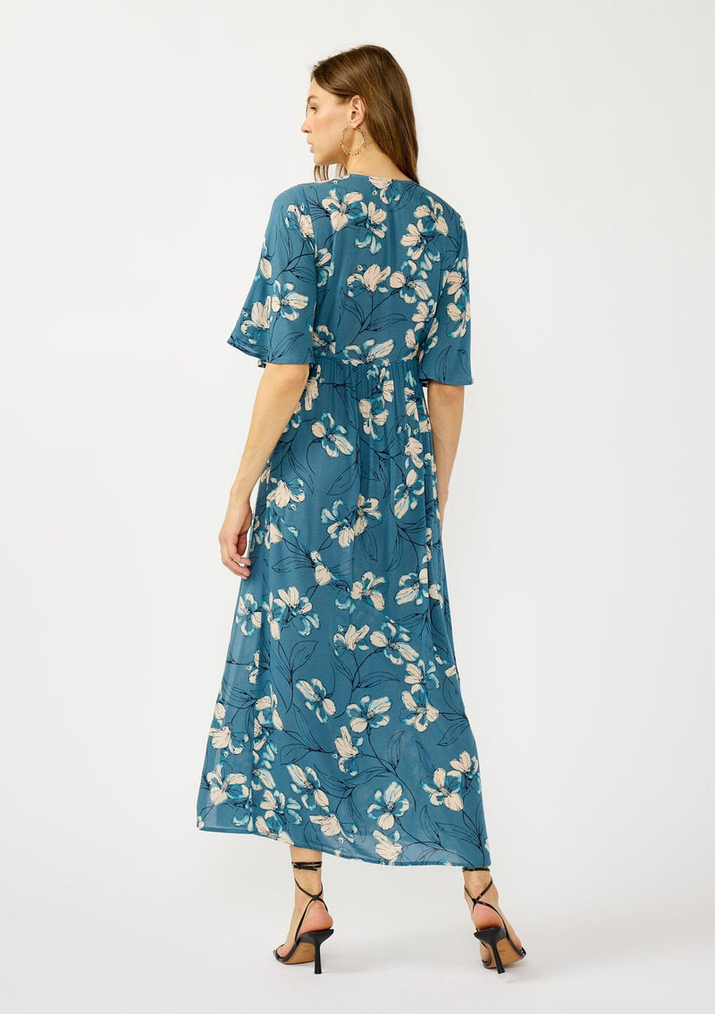 [Color: Teal/Blue] A back facing image of a blonde model wearing a teal floral print empire waist maxi dress with a deep v-neckline, short flutter sleeves, and a chic side slit. An effortless casual fall dress for special occasions. 