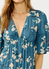 [Color: Teal/Blue] A detail image of a blonde model wearing a teal floral print empire waist maxi dress with a deep v-neckline, short flutter sleeves, and a chic side slit. An effortless casual fall dress for special occasions. 