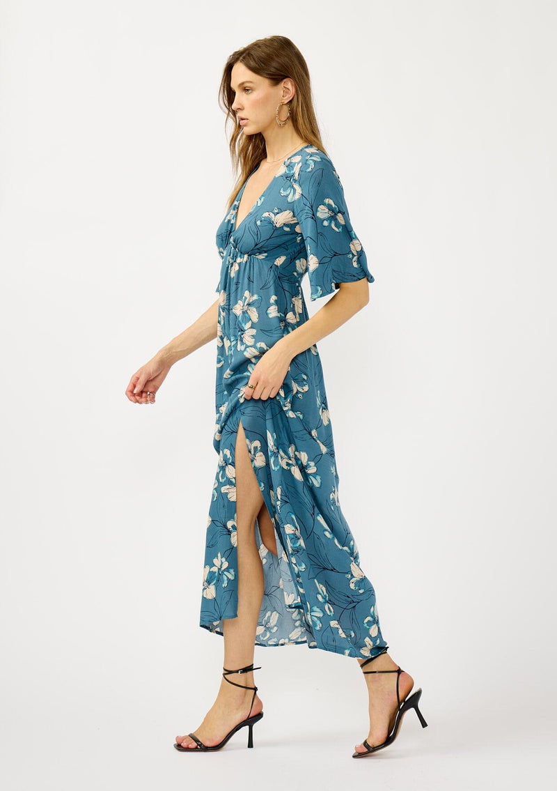 [Color: Teal/Blue] A side facing image of a blonde model wearing a teal floral print empire waist maxi dress with a deep v-neckline, short flutter sleeves, and a chic side slit. An effortless casual fall dress for special occasions. 