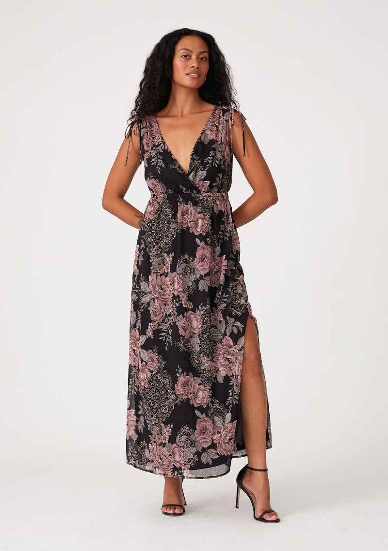[Color: Black/Dusty Rose] A front facing image of a brunette model wearing a bohemian fall mid length dress designed in a black and pink floral print, with metallic details throughout. With a surplice v neckline, an elastic empire waist, a side slit, and thick tank top straps with tie shoulder details. 