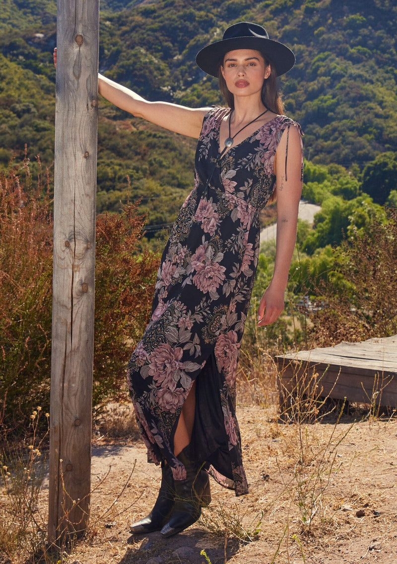 [Color: Black/Dusty Rose] A front facing image of a brunette model standing outside wearing a bohemian fall mid length dress designed in a black and pink floral print, with metallic details throughout. With a surplice v neckline, an elastic empire waist, a side slit, and thick tank top straps with tie shoulder details.