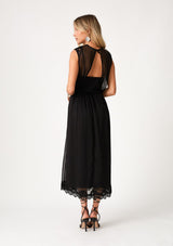 [Color: Black] A back facing image of a blonde model wearing a black bohemian holiday mid length special occasion dress. A sleeveless holiday dress designed in chiffon, with lace trim, a surplice v neckline, an empire waist, a half smocked bodice at the back, and an open back detail with single button closure. 