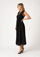 [Color: Black] A side facing image of a blonde model wearing a black bohemian holiday mid length special occasion dress. A sleeveless holiday dress designed in chiffon, with lace trim, a surplice v neckline, an empire waist, a half smocked bodice at the back, and an open back detail with single button closure. 