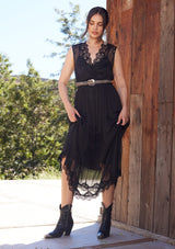 [Color: Black] A front facing image of a brunette model standing outside wearing a black bohemian holiday mid length special occasion dress. A sleeveless holiday dress designed in chiffon, with lace trim, a surplice v neckline, an empire waist, a half smocked bodice at the back, and an open back detail with single button closure.