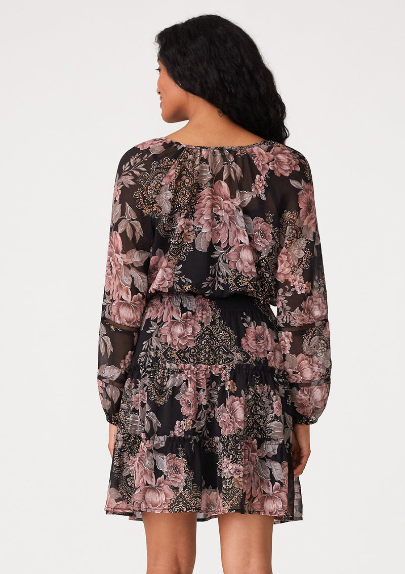 [Color: Black/Dusty Rose] A back facing image of a brunette model wearing a bohemian fall mini dress in a black and pink floral print. With voluminous long sleeves, a split v neckline with ties, a tiered mini skirt, a smocked elastic waist, and sheer mesh details. 
