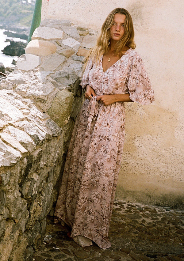 [Color: Dusty Rose/Light Brown] A front facing image of a blonde model wearing a bohemian pink floral maxi dress with short flutter sleeves, a v neckline, a long flowy skirt, and a self tie waist belt.