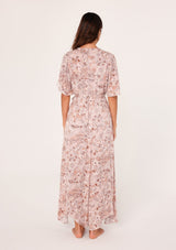 [Color: Dusty Rose/Light Brown] A back facing image of a brunette model wearing a bohemian pink floral maxi dress with short flutter sleeves, a v neckline, a long flowy skirt, and a self tie waist belt. 