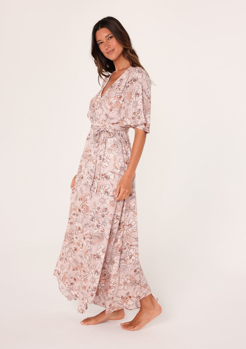 [Color: Dusty Rose/Light Brown] A side facing image of a brunette model wearing a bohemian pink floral maxi dress with short flutter sleeves, a v neckline, a long flowy skirt, and a self tie waist belt. 