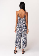 [Color: Navy/Natural] A back facing image of a brunette model wearing a sleeveless blue floral one piece jumpsuit. With adjustable spaghetti straps, a scoop neckline, a cropped wide leg, and a loose, relaxed fit. 