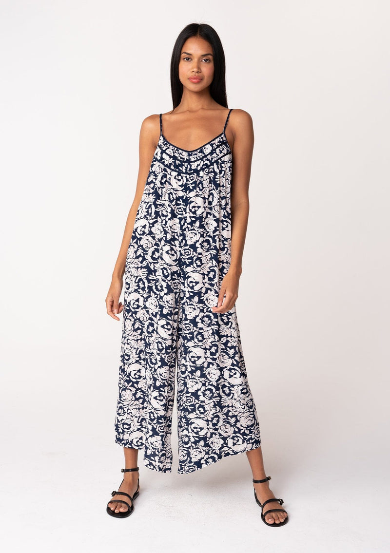 [Color: Navy/Natural] A front facing image of a brunette model wearing a sleeveless blue floral one piece jumpsuit. With adjustable spaghetti straps, a scoop neckline, a cropped wide leg, and a loose, relaxed fit. 