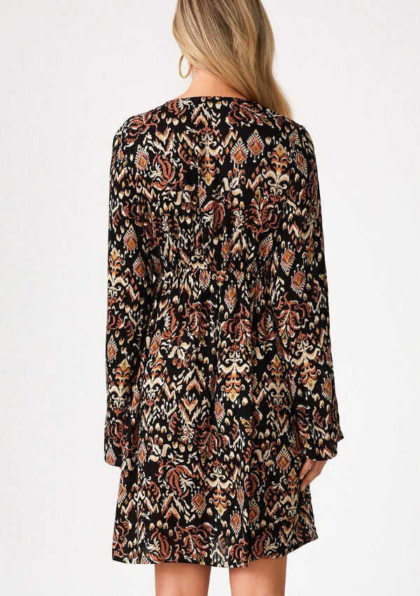 [Color: Black/Taupe] A back facing image of a blonde model wearing a bohemian fall mini dress in a bohemian brown print. With an empire waist, a deep v neckline, a relaxed flowy fit, and long bell sleeves with a split wrist detail. 