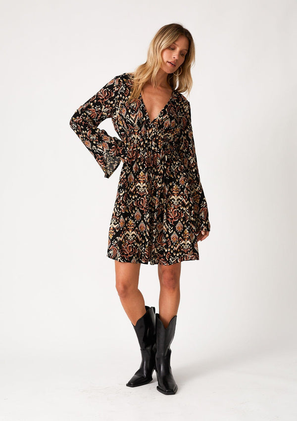 [Color: Black/Taupe] A front facing image of a blonde model wearing a bohemian fall mini dress in a bohemian brown print. With an empire waist, a deep v neckline, a relaxed flowy fit, and long bell sleeves with a split wrist detail. 