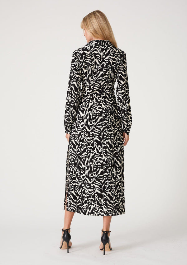 [Color: Black/Natural] A back facing image of a blonde model wearing a bohemian resort maxi shirt dress in a black and off white abstract print. With a self covered button front, long sleeves, a collared neckline, side slits, and an attached waist tie. 