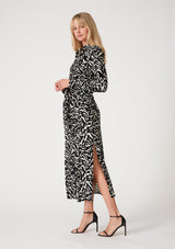[Color: Black/Natural] A side facing image of a blonde model wearing a bohemian resort maxi shirt dress in a black and off white abstract print. With a self covered button front, long sleeves, a collared neckline, side slits, and an attached waist tie. 