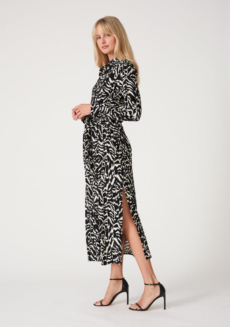 [Color: Black/Natural] A side facing image of a blonde model wearing a bohemian resort maxi shirt dress in a black and off white abstract print. With a self covered button front, long sleeves, a collared neckline, side slits, and an attached waist tie. 