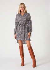 [Color: Grey/Cream] A full body front facing image of a blonde model wearing a relaxed fit mini shirt dress in an abstract grey print. With long sleeves, a gathered drawstring sleeve detail, tie wrist cuffs, a high low hemline, a self covered button front, side pockets, a collared neckline, and a tie waist belt. 