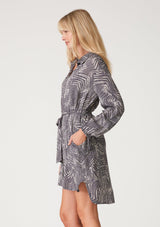 [Color: Grey/Cream] A half body side facing image of a blonde model wearing a relaxed fit mini shirt dress in an abstract grey print. With long sleeves, a gathered drawstring sleeve detail, tie wrist cuffs, a high low hemline, a self covered button front, side pockets, a collared neckline, and a tie waist belt. 