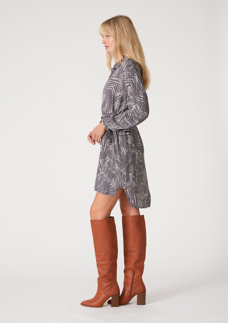[Color: Grey/Cream] A side facing image of a blonde model wearing a relaxed fit mini shirt dress in an abstract grey print. With long sleeves, a gathered drawstring sleeve detail, tie wrist cuffs, a high low hemline, a self covered button front, side pockets, a collared neckline, and a tie waist belt. 