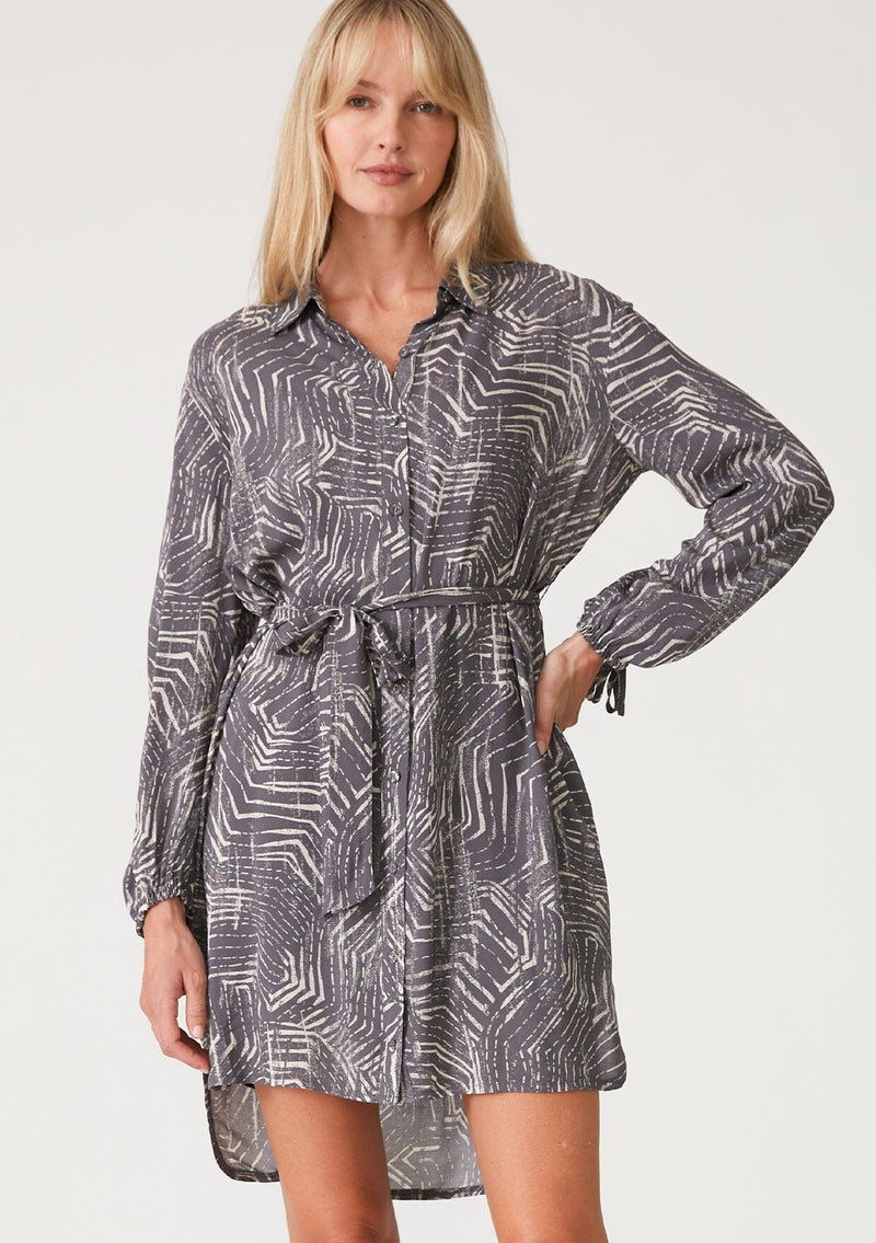 [Color: Grey/Cream] A front facing image of a blonde model wearing a relaxed fit mini shirt dress in an abstract grey print. With long sleeves, a gathered drawstring sleeve detail, tie wrist cuffs, a high low hemline, a self covered button front, side pockets, a collared neckline, and a tie waist belt. 