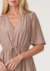 [Color: Mocha] A close up front facing image of a blonde model wearing a mocha brown bohemian resort mini dress. With short flutter sleeves, a v neckline, an adjustable drawstring tie waist, and a mini pom trim. 
