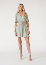 [Color: Pistachio] A front facing image of a blonde model wearing a light green bohemian resort mini dress. With short flutter sleeves, a v neckline, an adjustable drawstring tie waist, and a mini pom trim. 