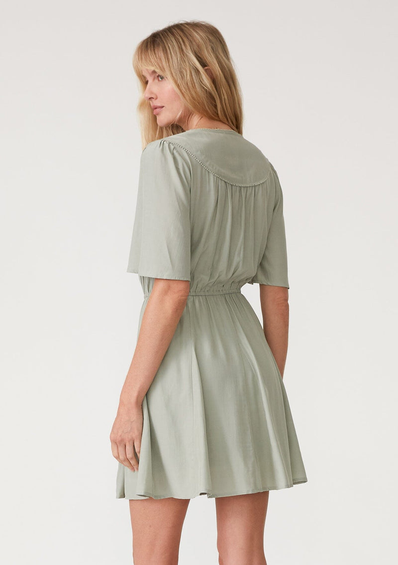 [Color: Pistachio] A back facing image of a blonde model wearing a light green bohemian resort mini dress. With short flutter sleeves, a v neckline, an adjustable drawstring tie waist, and a mini pom trim. 