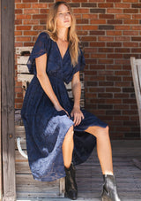 [Color: Navy] A sitting image of a blonde model wearing a blue floral jacquard maxi dress with gold foil accents. Featuring a flattering surplice v-neckline, dolman sleeves, elastic waist, tie back, and a handkerchief hemline. 