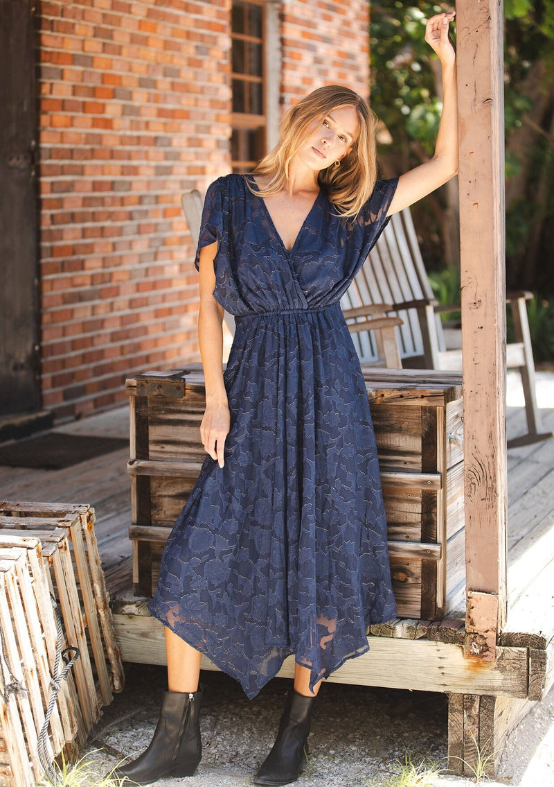 [Color: Navy] A front facing image of a blonde model wearing a blue floral jacquard maxi dress with gold foil accents. Featuring a flattering surplice v-neckline, dolman sleeves, elastic waist, tie back, and a handkerchief hemline. 