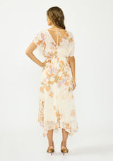 [Color: Ivory/Rust] A back facing image of a brunette model wearing an ivory and orange floral dress with a surplice v neckline, short dolman sleeves, an open tie back detail, and an asymmetrical hem. 