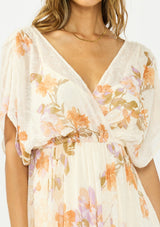 [Color: Ivory/Rust] A detail image of a brunette model wearing an ivory and orange floral dress with a surplice v neckline, short dolman sleeves, an open tie back detail, and an asymmetrical hem. 