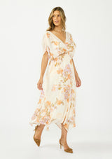 [Color: Ivory/Rust] A side facing image of a brunette model wearing an ivory and orange floral dress with a surplice v neckline, short dolman sleeves, an open tie back detail, and an asymmetrical hem. 