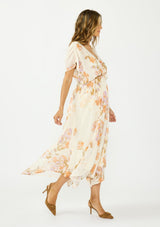 [Color: Ivory/Rust] A side facing image of a brunette model wearing an ivory and orange floral dress with a surplice v neckline, short dolman sleeves, an open tie back detail, and an asymmetrical hem. 