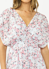 [Color: Grey/Dusty Rose] A detail image of a brunette model wearing a floral maxi dress with a burnout texture, short dolman sleeves, an asymmetrical hem, and a tie back detail. 
