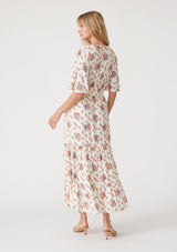 [Color: Natural/Rust] A back facing image of a blonde model wearing a classic bohemian maxi dress in an off white and pink floral print. With short flutter sleeves, a surplice v neckline, a smocked elastic waist, a drawstring tie waist, and a long flowy tiered skirt. 