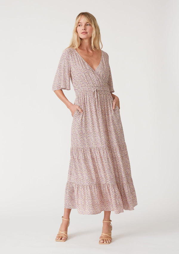 [Color: Ivory/Purple] A front facing image of a blonde model wearing a classic bohemian maxi dress in an off white and purple ditsy floral print. With short flutter sleeves, a surplice v neckline, a smocked elastic waist, a drawstring tie waist, and a long flowy tiered skirt.