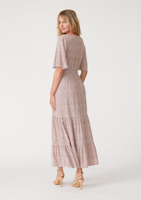 [Color: Ivory/Purple] A back facing image of a blonde model wearing a classic bohemian maxi dress in an off white and purple ditsy floral print. With short flutter sleeves, a surplice v neckline, a smocked elastic waist, a drawstring tie waist, and a long flowy tiered skirt.
