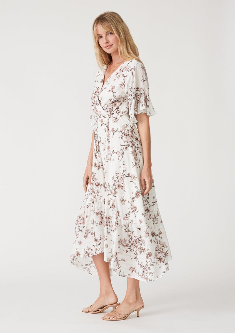 [Color: Ivory/Taupe] A side facing image of a blonde model wearing an ivory and taupe floral print bohemian maxi dress with sparkly metallic thread detail. With short flutter sleeves, a long tiered skirt, a surplice v neckline, and an adjustable drawstring waist. 
