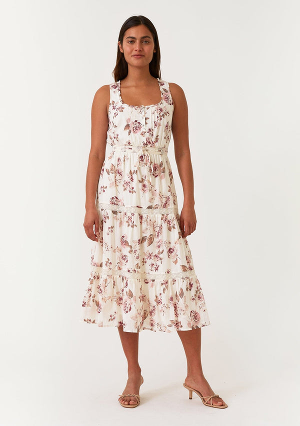 [Color: Natural/Dusty Wine] A front facing image of a brunette model wearing a sleeveless bohemian mid length dress in an off white and pink floral print. With a ruffle trimmed scoop neckline, a self covered button front top, a tiered skirt, lace trim, pintuck details, and a drawstring waist with ties. 